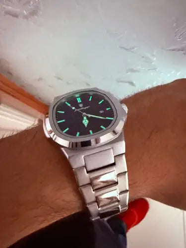 A sleek silver-toned stainless steel watch with a black dial and luminous green hour markers, featuring roman numerals at the 12 o’clock position.
